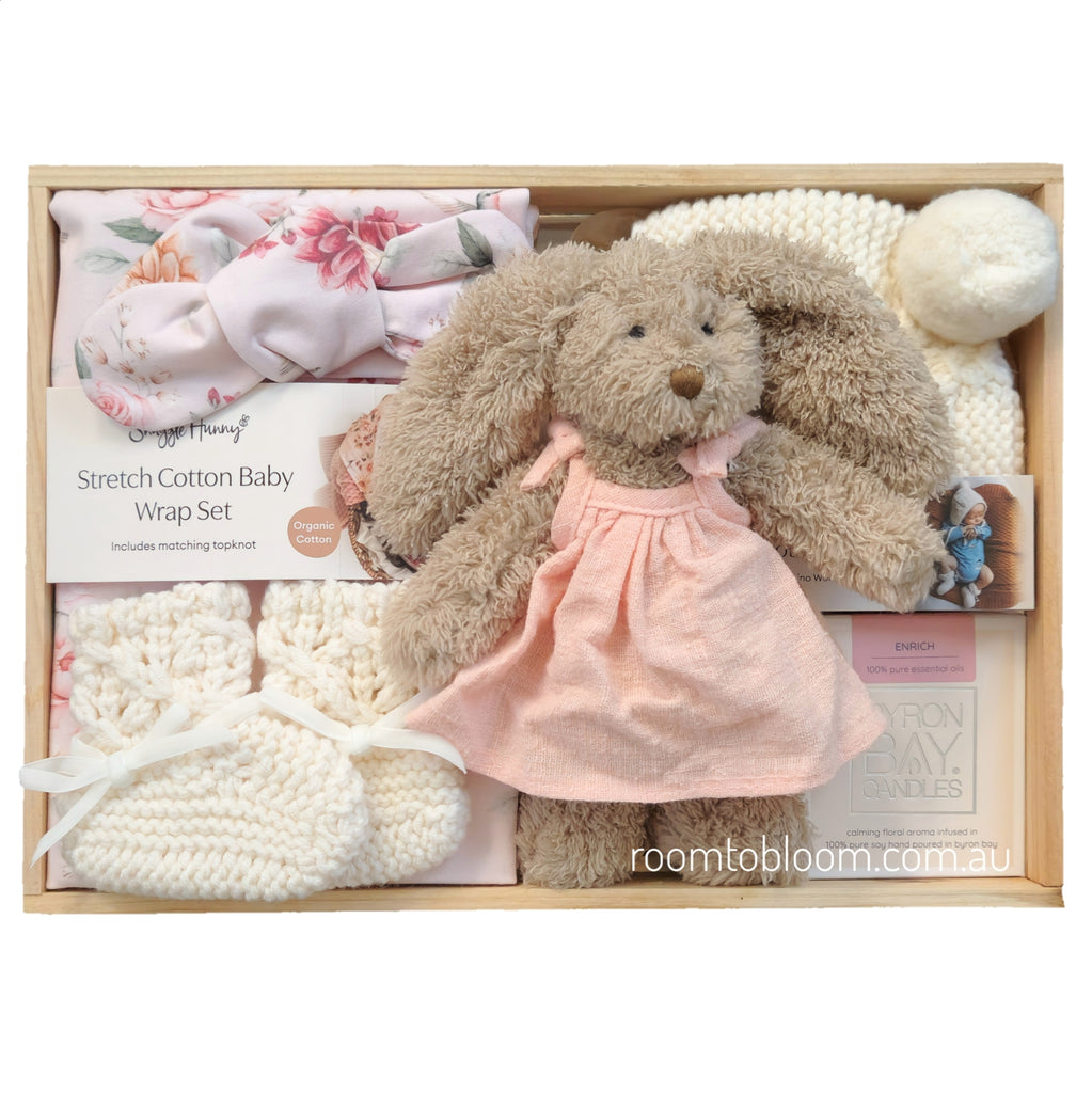 ROOM TO BLOOM Bambina Baby Gift Hamper set in a wooden box featuring a plush brown bunny doll in a pink dress, a pink floral wrap, cream knit socks, a cream knit hat, and more.
