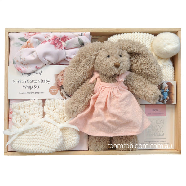ROOM TO BLOOM Bambina Baby Gift Hamper set in a wooden box featuring a plush brown bunny doll in a pink dress, a pink floral wrap, cream knit socks, a cream knit hat, and more.