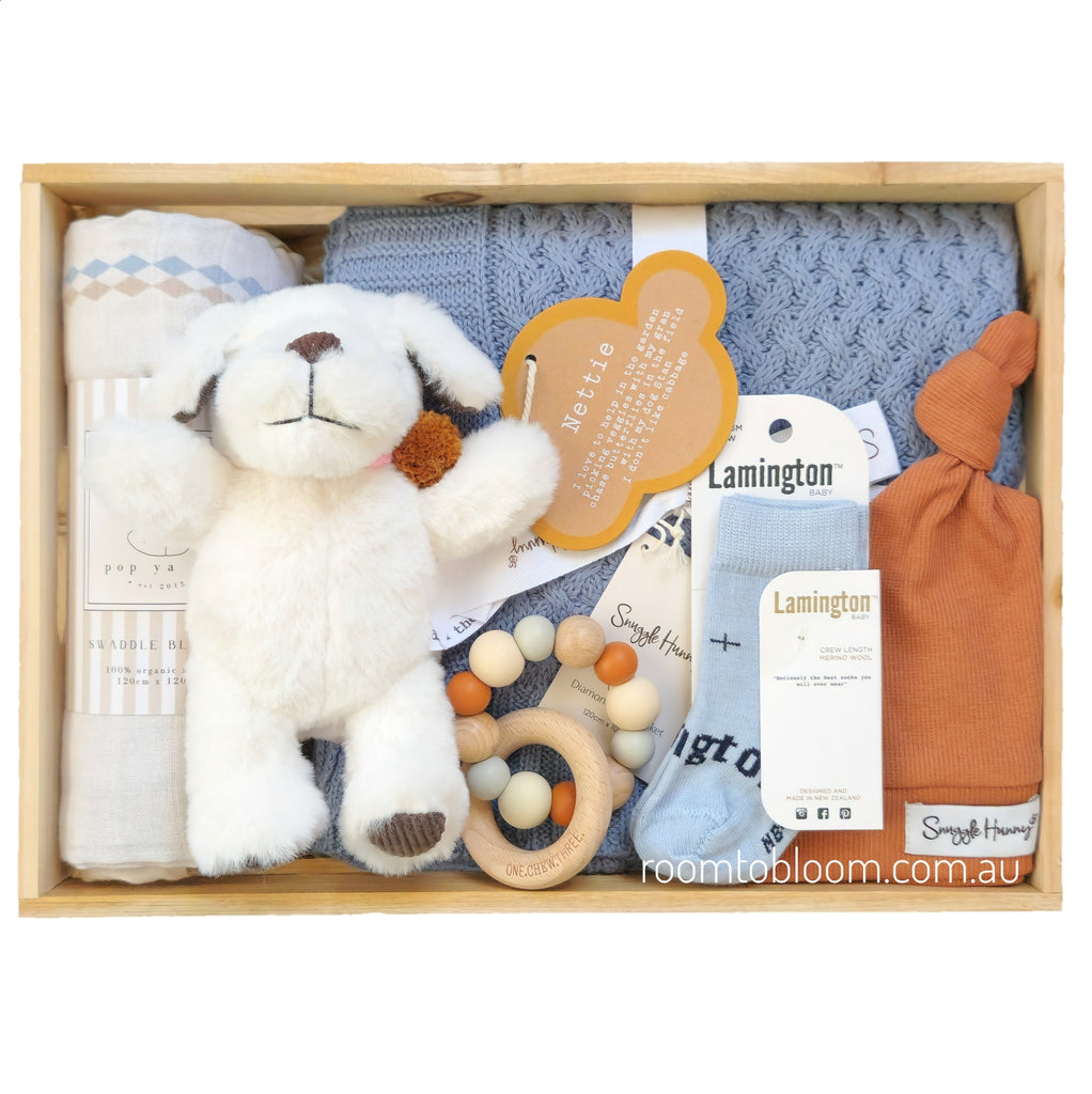 ROOM TO BLOOM Barkley Baby Gift Hamper set arranged in a wooden box, featuring a white plush dog toy, a blue knit blanket, a white printed swaddle, light blue socks, a terracotta baby beanie, and a wooden bead teether.