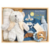 ROOM TO BLOOM Bear Hugs Baby Gift Hamper set in a wooden box featuring a plush grey teddy bear with a cream knit jacket, a folded muslin wrap, a blue baby playsuit with a croissant applique, a blue book titled 'On The Night You Were Born' by Nancy Tillman, a light blue hat, and a wooden teething ring.