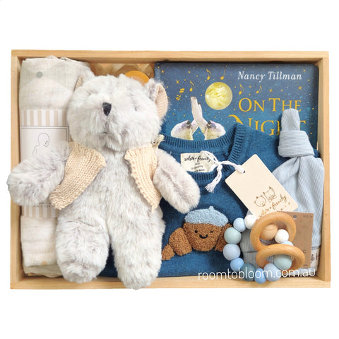 ROOM TO BLOOM Bear Hugs Baby Gift Hamper set in a wooden box featuring a plush grey teddy bear with a cream knit jacket, a folded muslin wrap, a blue baby playsuit with a croissant applique, a blue book titled 'On The Night You Were Born' by Nancy Tillman, a light blue hat, and a wooden teething ring.