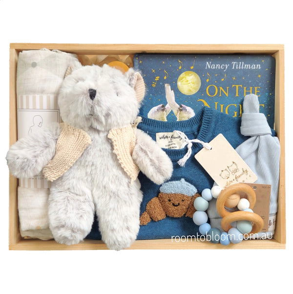 ROOM TO BLOOM Bear Hugs Baby Gift Hamper set in a wooden box featuring a plush grey teddy bear with a cream knit jacket, a folded muslin wrap, a blue baby playsuit with a croissant applique, a blue book titled 'On The Night You Were Born' by Nancy Tillman, a light blue hat, and a wooden teething ring.