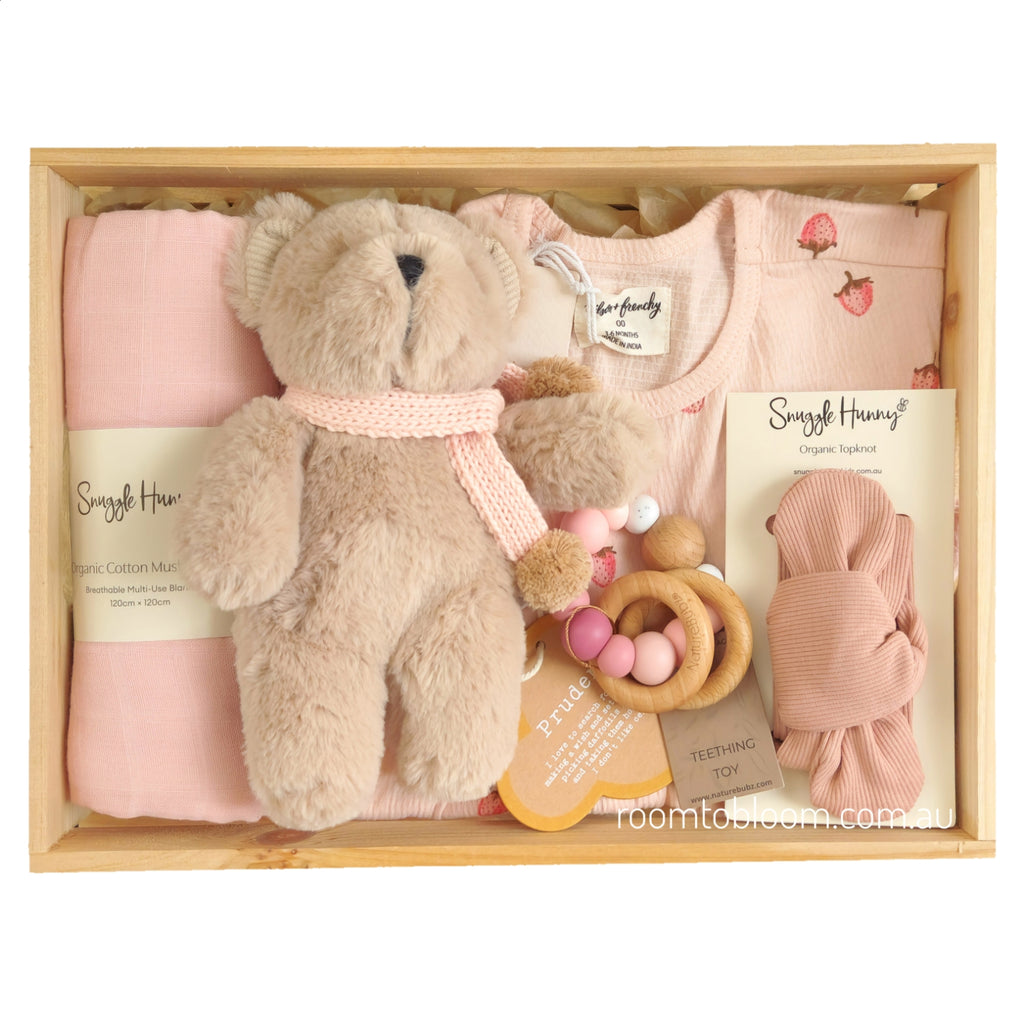 ROOM TO BLOOM Berry Bliss Baby Gift Hamper set arranged in a light wood box, featuring a pink short-sleeve dress with strawberry print, a folded pink swaddle, a plush teddy bear wearing a pink knit scarf, a pink teething toy, and a pink headband.