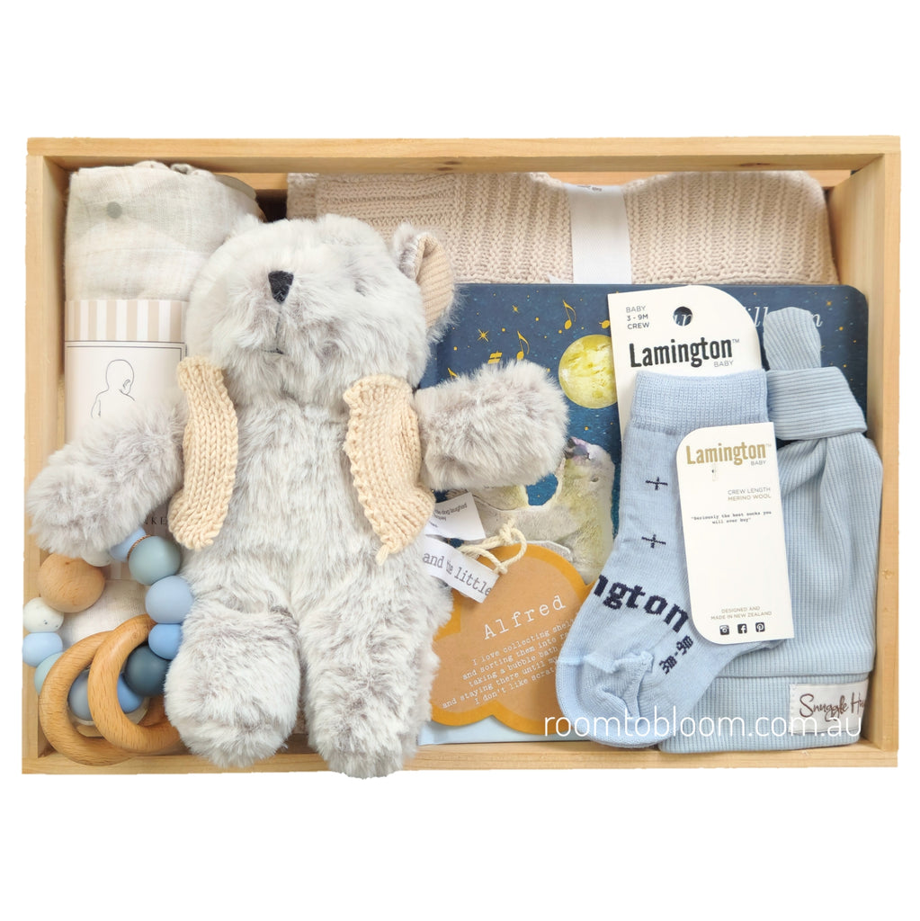 ROOM TO BLOOM Big Dreams Baby Gift Hamper set arranged in a light wood box, featuring a plush grey stuffed bear, light blue knit baby socks, a grey knit blanket, wooden and blue beads, a patterned muslin swaddle, a beanie and a picture book. 