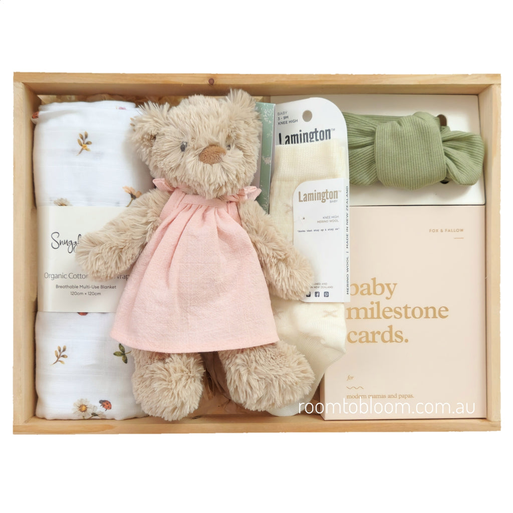 ROOM TO BLOOM Blessings Baby Gift Hamper set in a light wood box featuring a plush teddy bear wearing a pink dress, folded white swaddle with floral embroidery, a green bow, Lamington socks, and baby milestone cards.
