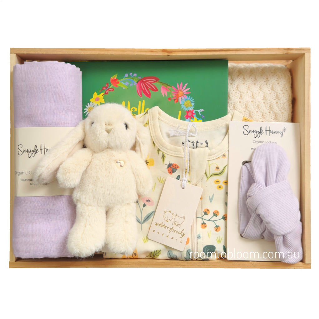 ROOM TO BLOOM Bloom Box Baby Gift Hamper set in a wooden box, featuring a cream plush bunny, a cream floral baby zipsuit, a light purple muslin blanket, a cream blamket, a picture book, and a light purple handband, arranged neatly inside the box.