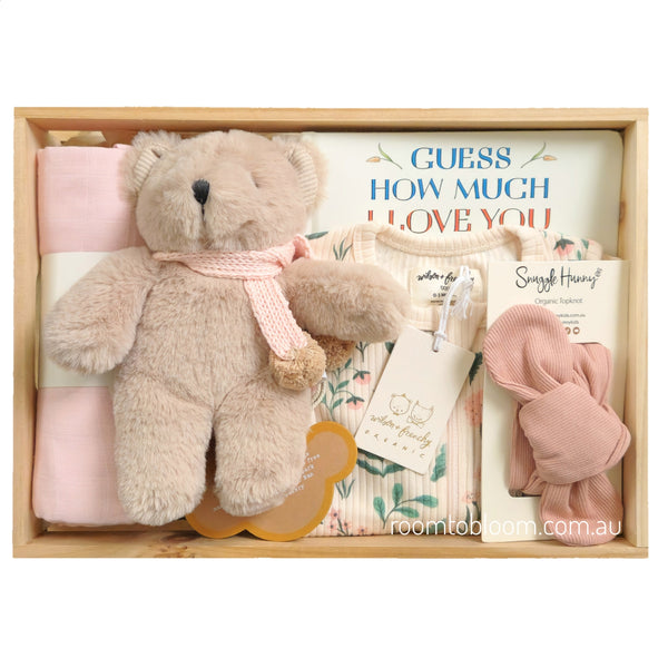 ROOM TO BLOOM Blooming Beauty Baby Gift Hamper set in a light wood box featuring a plush teddy bear with a pink scarf, a folded pink swaddle, a floral baby outfit, a pink bow, and a book titled 'GUESS HOW MUCH I LOVE YOU'.