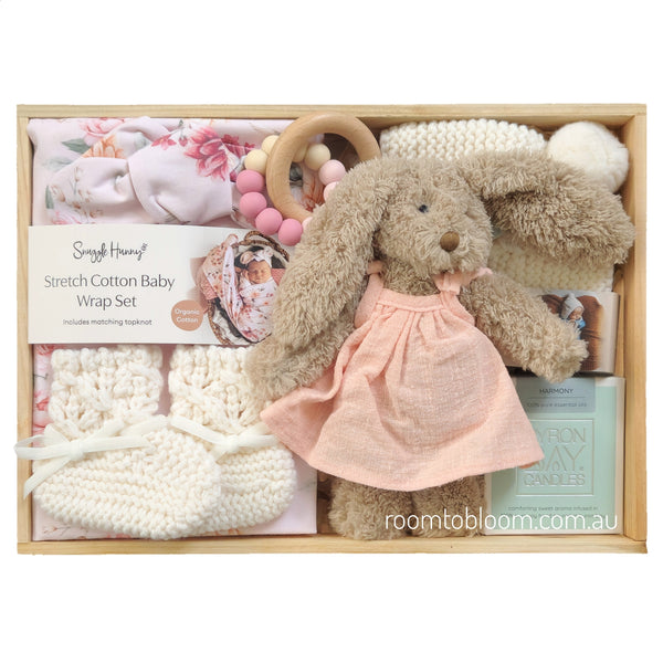 ROOM TO BLOOM Blooms Baby Gift Hamper set in a light wood box from featuring a stretch cotton wrap set, a plush bunny doll in a pink dress, a wooden ring teether with pink and cream beads, cream knit booties and bonnet, and a candle.