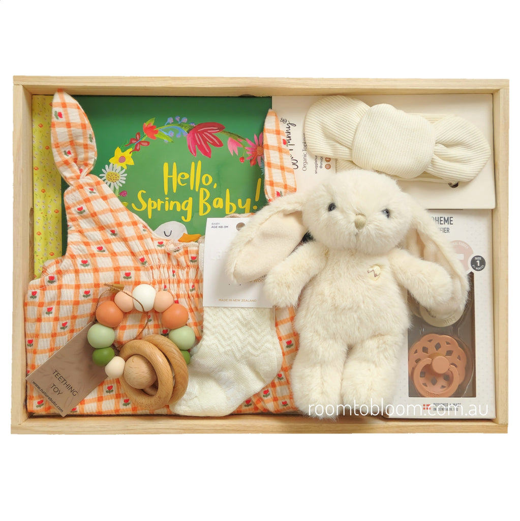 ROOM TO BLOOM Blossoming Baby Gift Hamper set arranged in a light wood tray, featuring a cream plush bunny, an orange-and-white gingham baby playsuit, cream knit socks, a wooden teething toy, a green 'Hello, Spring Baby!' book, a cream fabric headband, and a set of two pacifiers.