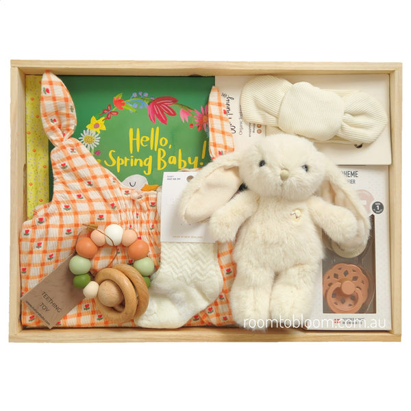 ROOM TO BLOOM Blossoming Baby Gift Hamper set arranged in a light wood tray, featuring a cream plush bunny, an orange-and-white gingham baby playsuit, cream knit socks, a wooden teething toy, a green 'Hello, Spring Baby!' book, a cream fabric headband, and a set of two pacifiers.