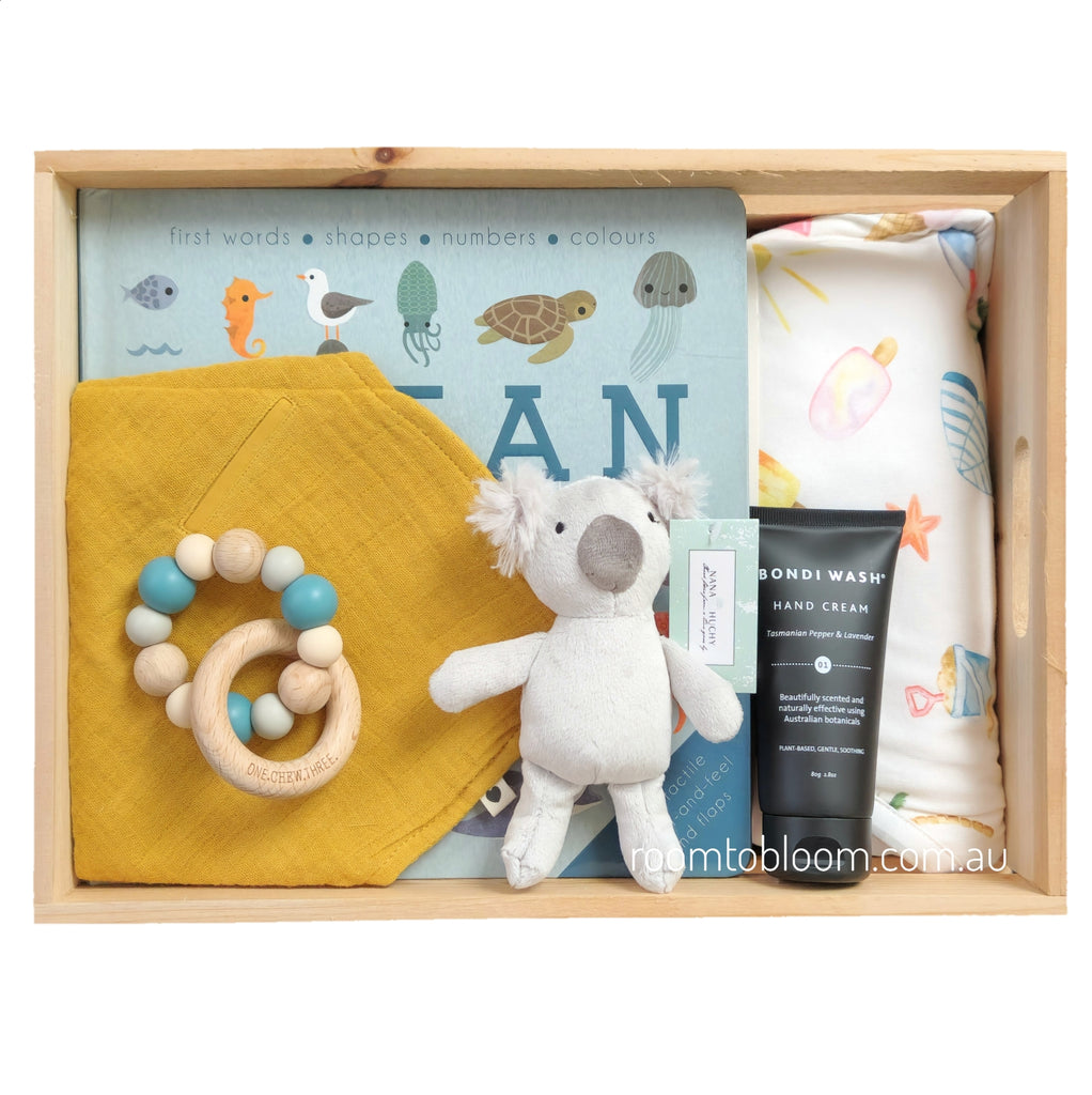 ROOM TO BLOOM Bondi Baby Gift Hamper set arranged in a light wood tray, including a blue children's book with ocean creatures, a yellow muslin bib, a wooden bead teether, a greykoala plush toy, and a black Bondi Wash hand cream.