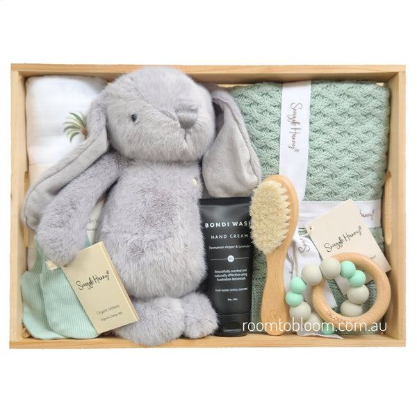 ROOM TO BLOOM Calm Palm Baby Gift Hamper set arranged in a light wood box, featuring a plush grey bunny toy, a mint green textured blanket, a folded white swaddle, a black hand cream tube, a wooden baby brush, a wooden teething ring with mint and white beads, and sage mittens. 