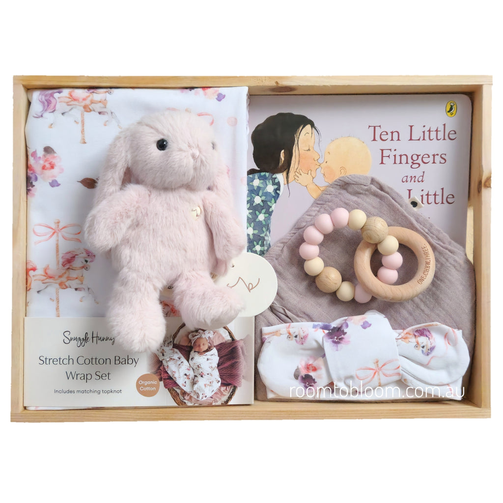 ROOM TO BLOOM Carousel Baby Gift Hamper set arranged inside a light wood box. The set includes a pink plush bunny, a floral stretch cotton baby wrap and headband set, a wooden bead teether, a bandana bib, and a baby book titled 'Ten Little Fingers and Ten Little Toes'.