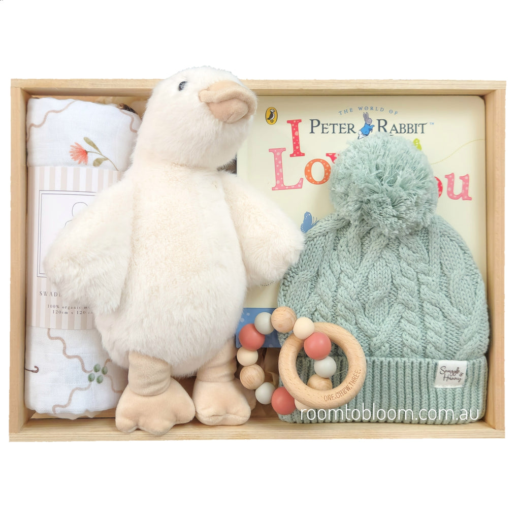 ROOM TO BLOOM Chelsea Baby Gift Hamper set in a light wood box featuring a plush cream goose toy, a mint green cable-knit pom-pom hat, a wooden bead teething ring, a folded patterned swaddle, and a Peter Rabbit book.