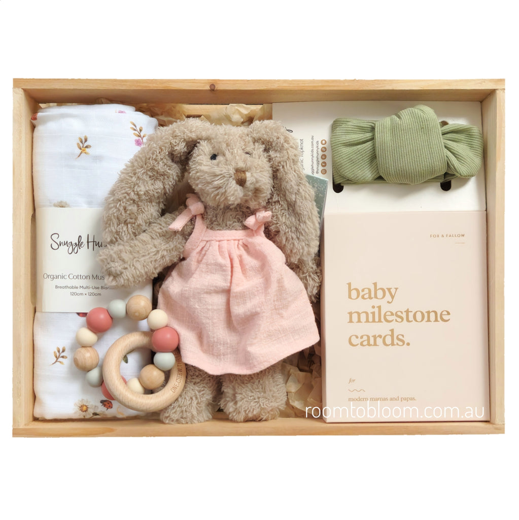 ROOM TO BLOOM Cherished Baby Gift Hamper set arranged in a wooden box, featuring a plush bunny doll in a pink dress, white cotton muslin swaddle, a sage green ribbed bow headband, wooden beaded teether, and a milestone card set.