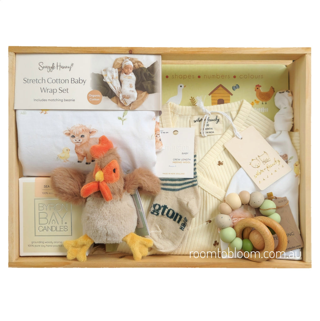 ROOM TO BLOOM Clucky Charm Baby Gift Hamper set arranged in a light wood box, including a Stretch Cotton Baby Wrap Set, a plush rooster toy, a wooden teething ring, and other baby items.