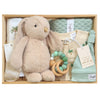 ROOM TO BLOOM Copacabana Baby Gift Hamper set in a light wood box featuring a plush beige bunny toy, a printed cotton romper and swaddle, a mint green knit blanket, a wooden ring teether with pastel beads, a beanie and socks.