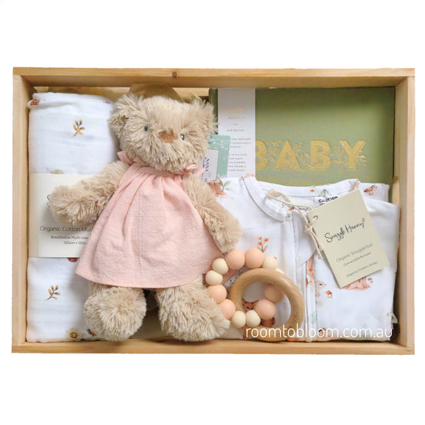 ROOM TO BLOOM Forever Friends Baby Gift Hamper set arranged inside a light wood box on a white background. The set includes a beige teddy bear wearing a pale pink dress, folded white baby clothes, a wooden bead teether, a green baby book with gold lettering, and a swaddle.