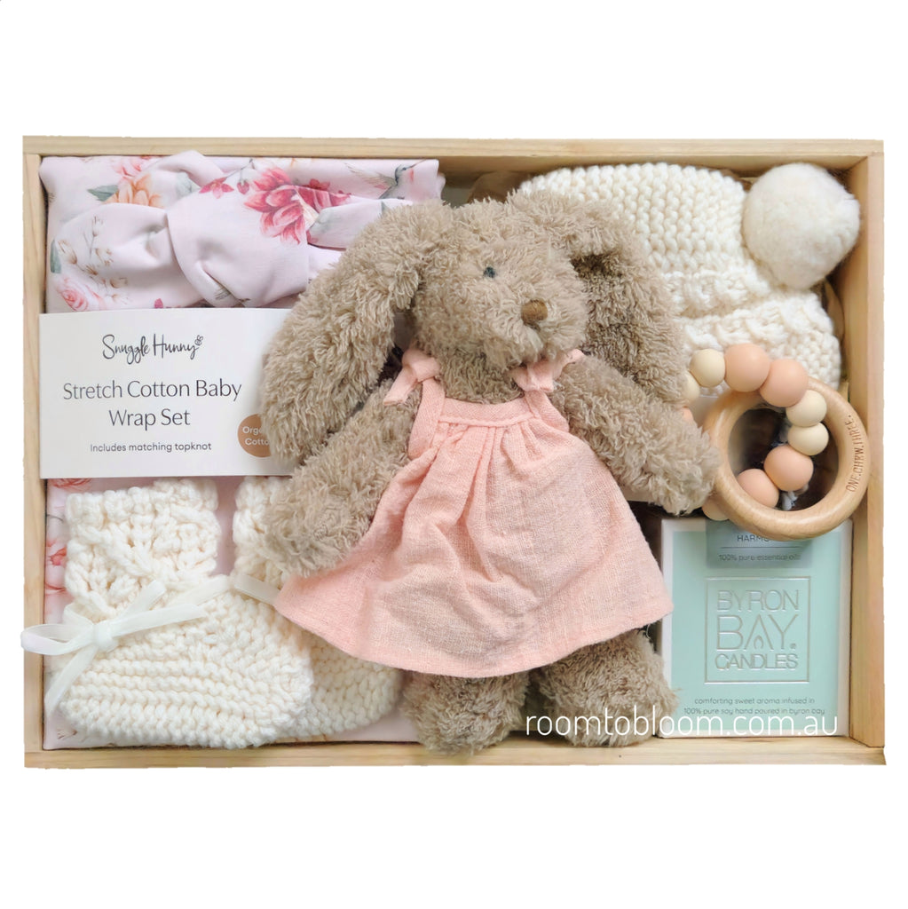 ROOM TO BLOOM Harmony Baby Gift Hamper set in a light wood box, featuring a plush brown bunny doll in a pink dress, a pink floral baby wrap and headband set, white knit booties and bonnet, a wooden teething ring, and a candle.
