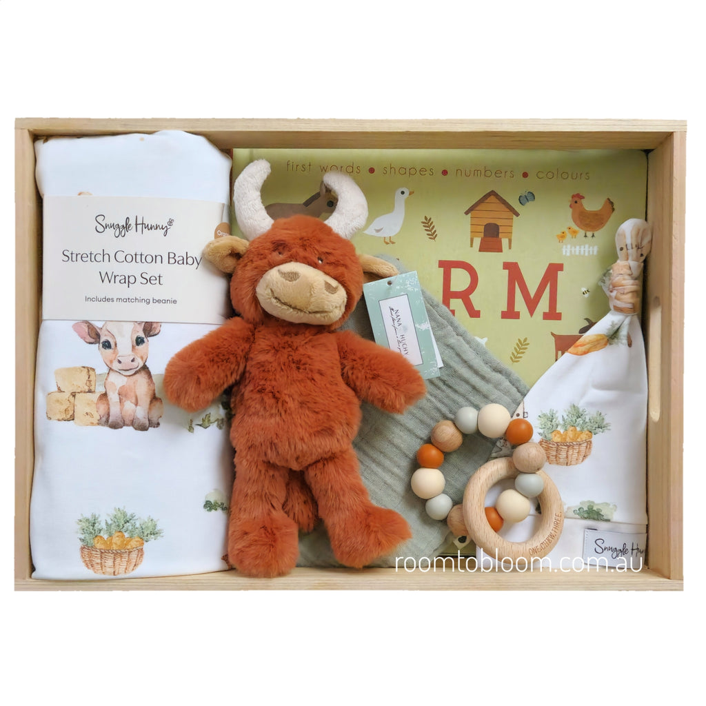 ROOM TO BLOOM Highlander Baby Gift Hamper set in a wooden box featuring a white stretch cotton baby wrap ad beanie set with farm illustrations, a plush brown bull toy, a green farm-themed book, a wooden bead rattle, and a sage bb.