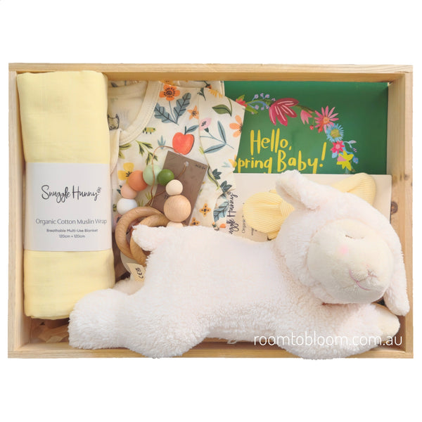 ROOM TO BLOOM Hope Baby Gift Hamper set arranged in a light wood box, featuring a white plush lamb toy, a folded pale yellow muslin wrap, a floral baby outfit, a book, a teether and a yellow headband.