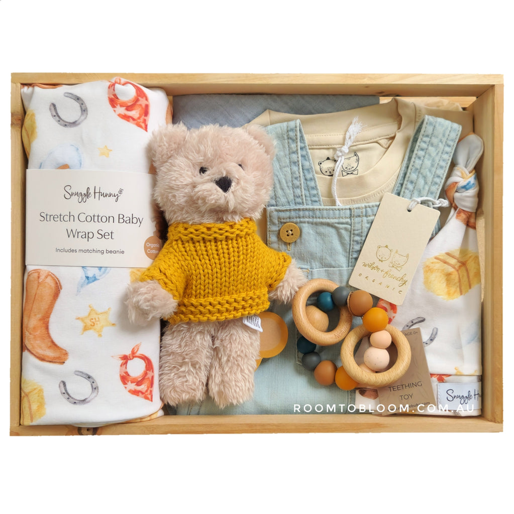 ROOM TO BLOOM Howdy Baby Gift Hamper set in a wooden box, featuring a plush teddy bear in a yellow knit sweater, a light blue denim overall and tee, a stretch cotton wrap and beanie set with cowboy print, wooden teething ring, and a bandana bib.