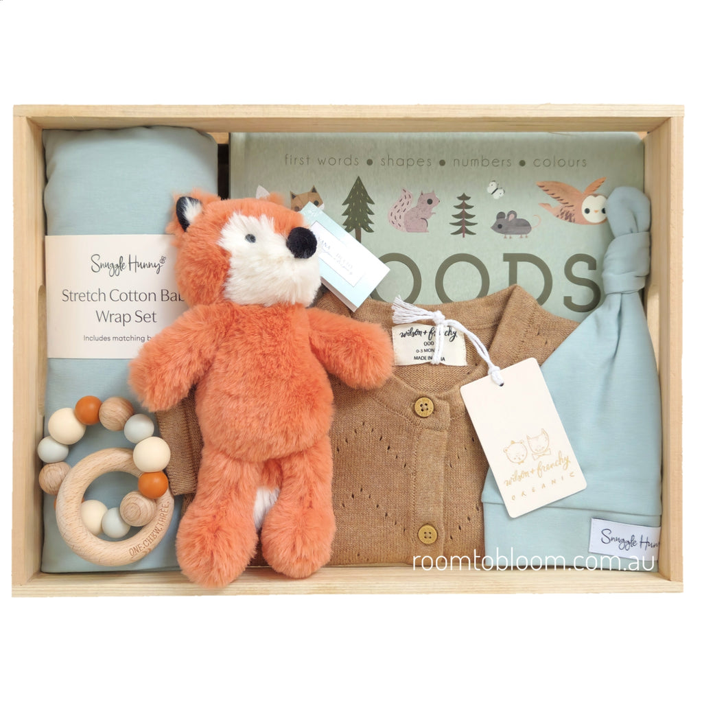 ROOM TO BLOOM In the Woods Baby Gift Hamper set in a light wood box featuring a lightg reenl stretch cotton blanket wrap set, a brown knitgrowsuit, a plush orange fox toy, a wooden ring teether, and a matching woodland-themed book.