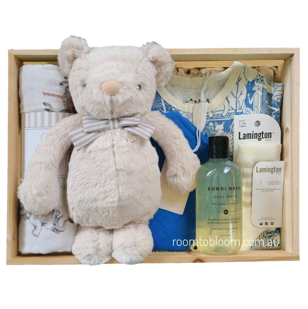 ROOM TO BLOOM Le Cirque Baby Gift Hamper set arranged in a light wood box, featuring a cream plush teddy bear with a striped bow tie, a zipsuit, a bottle of Bondi Wash baby wash, and Lamington socks and more.