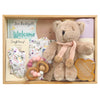ROOM TO BLOOM Little Flower Baby Gift Hamper set in a light wood box featuring a plush teddy bear with a pink knit pom-pom scarf, a floral baby headband, wooden teething ring, a floral zipsuit, a lavender muslin swaddle, and a book titled "Welcome to this World".