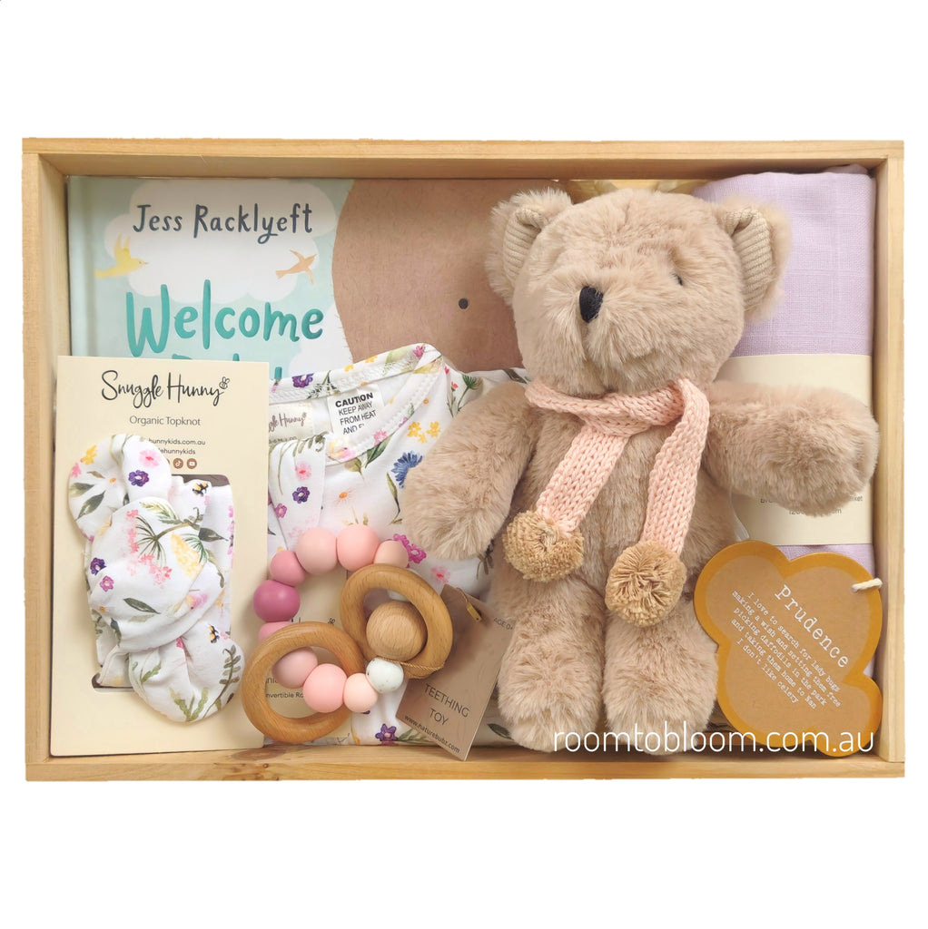 ROOM TO BLOOM Little Flower Baby Gift Hamper set in a light wood box featuring a plush teddy bear with a pink knit pom-pom scarf, a floral baby headband, wooden teething ring, a floral zipsuit, a lavender muslin swaddle, and a book titled "Welcome to this World".