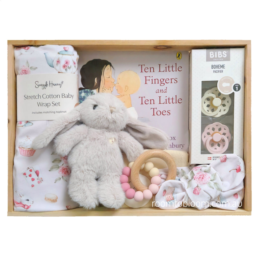 ROOM TO BLOOM Little Rabbit Baby Gift Hamper set arranged inside a wooden box, featuring a plush beige bunny toy, a pink and white floral baby wrap blanket and headband, a wooden ring teether with pink and cream beads, a book titled 'Ten Little Fingers and Ten Little Toes', and a boxed pacifier set.
