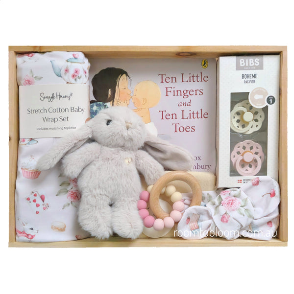 ROOM TO BLOOM Little Rabbit Baby Gift Hamper set arranged inside a wooden box, featuring a plush beige bunny toy, a pink and white floral baby wrap blanket and headband, a wooden ring teether with pink and cream beads, a book titled 'Ten Little Fingers and Ten Little Toes', and a boxed pacifier set.