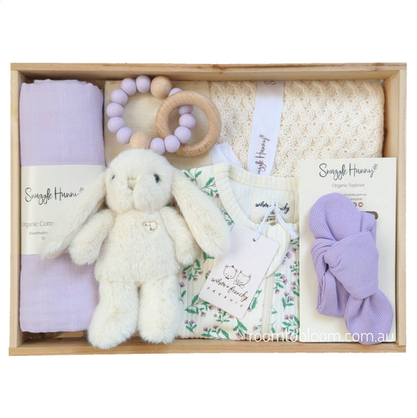 ROOM TO BLOOM Lovely Lilac Baby Gift Hamper set in a wooden box featuring a plush white bunny, a light purple cotton swaddle, a wooden bead teether with a lavender bead, a cream blanket, a floral baby romper, and a matching purple headband.