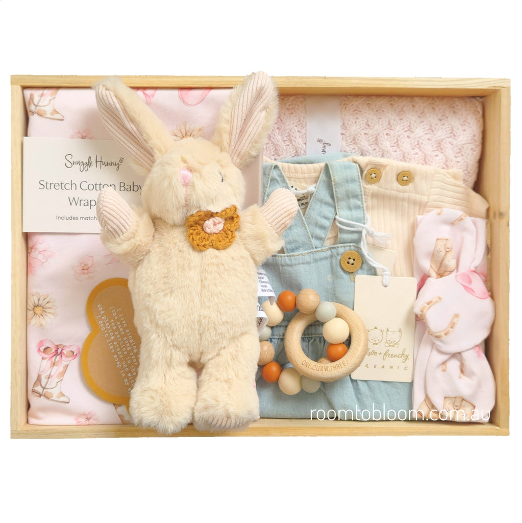 ROOM TO BLOOM Miss Daisy Baby Gift Hamper set in a wooden box featuring a cream plush bunny, a light blue denim baby romper, cream t-shirt, a pink knit blanket, a wooden bead teether, and a pink stretch baby wrap and headband. 