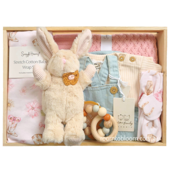 ROOM TO BLOOM Miss Dolly Baby Gift Hamper set arranged in a wooden box, featuring a cream plush bunny with a crocheted orange bow, a light blue denim baby outfit, a tee, a pink knit blanket, a floral stretch cotton baby wrap and matching headband, and a wooden bead teether