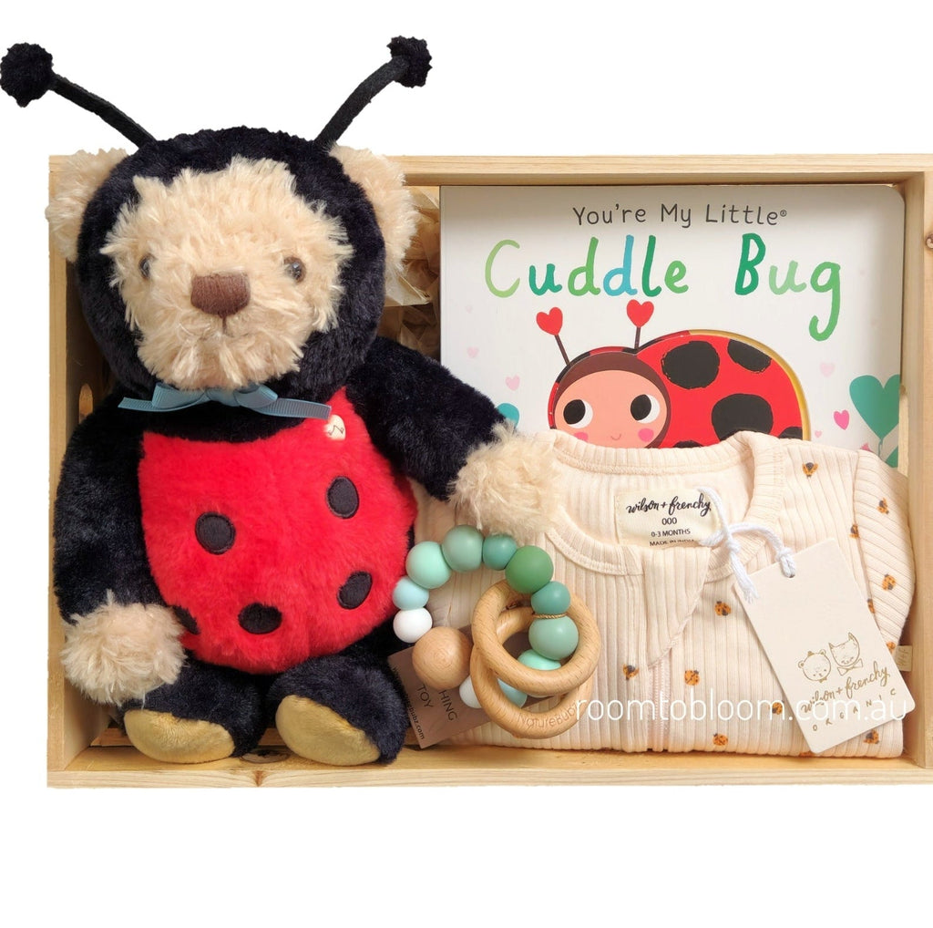 ROOM TO BLOOM My Little Cuddle Bug Baby Gift Hamper set arranged in a light wood box, featuring a plush ladybug toy, a cream ribbed baby bodysuit with a clover print, a wooden bead teether, and a children's book titled 'You're My Little Cuddle Bug'.