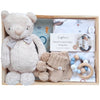 ROOM TO BLOOM Nautica Baby Gift Hamper set arranged inside a light wood tray on a white background. The set includes a beige plush teddy bear with a striped bow tie, knitted baby booties, a wooden ring teether with blue and white beads, a book, and a printed wrap and beanie.