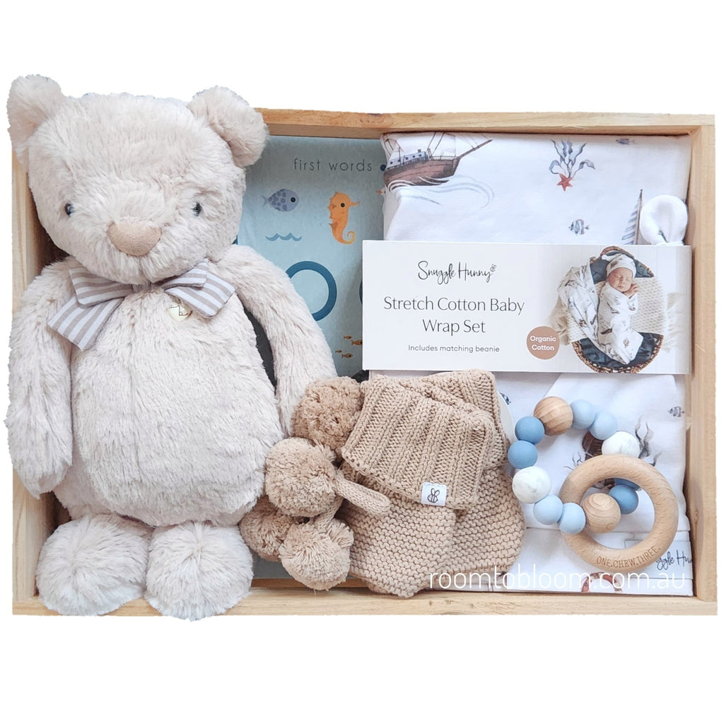 ROOM TO BLOOM Nautica Baby Gift Hamper set arranged inside a light wood tray on a white background. The set includes a beige plush teddy bear with a striped bow tie, knitted baby booties, a wooden ring teether with blue and white beads, a book, and a printed wrap and beanie.