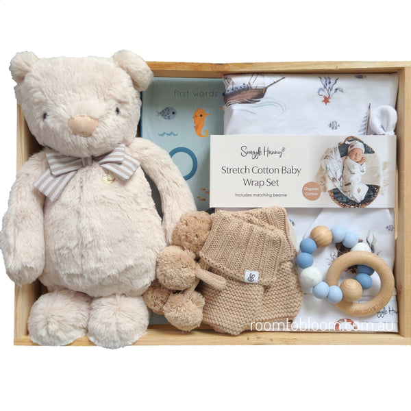 ROOM TO BLOOM Nautica Baby Gift Hamper set arranged inside a light wood tray on a white background. The set includes a beige plush teddy bear with a striped bow tie, knitted baby booties, a wooden ring teether with blue and white beads, a book, and a printed wrap and beanie.