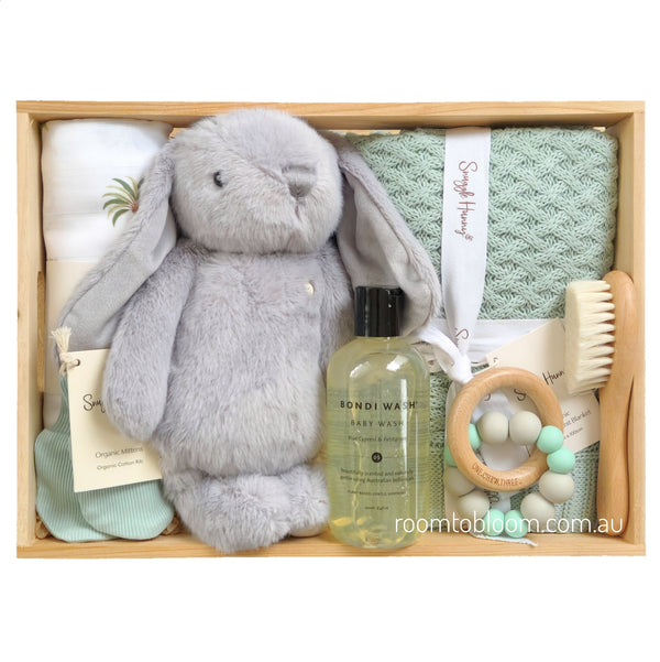 ROOM TO BLOOM Oasis Baby Gift Hamper set arranged in a light wood tray, featuring a plush grey bunny, a mint green textured towel, a bottle of Bondi Wash baby wash, a wooden teething ring with mint and white beads, a wooden baby brush, and folded muslin and sage baby blanket. 