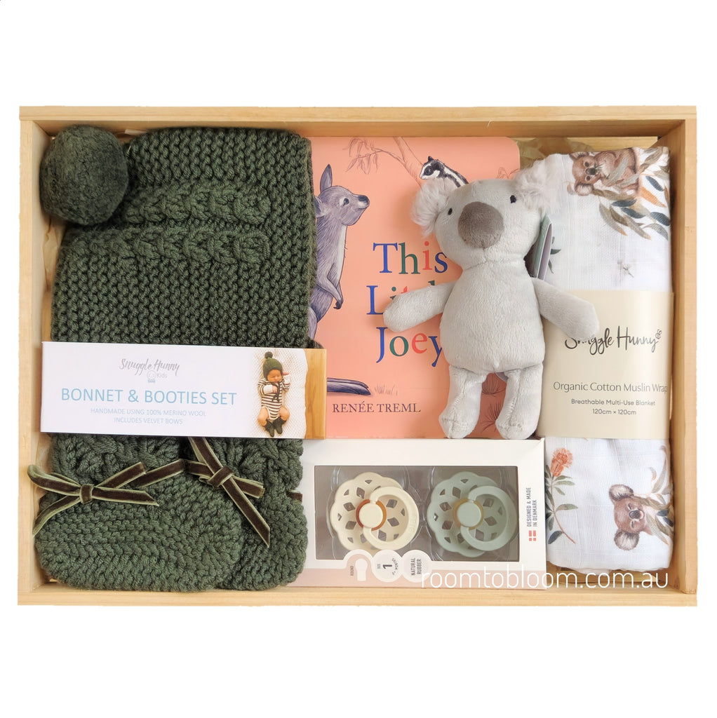 ROOM TO BLOOM One Little Joey Baby Gift Hamper set arranged in a wooden tray, featuring an olive green knit bonnet and bootie set, a plush koala toy, a book titled 'This Little Joey', a cotton muslin cloth, and pacifiers.