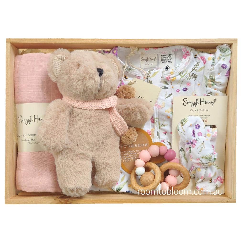 ROOM TO BLOOM Petite Fleur Baby Gift Hamper set in a wooden box featuring a plush teddy bear with a pink knit scarf, a folded pink swaddle, a floral baby bodysuit, a matching folded headband, and a wooden bead teether.