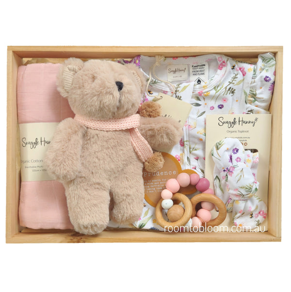 ROOM TO BLOOM Petite Fleur Baby Gift Hamper set in a wooden box featuring a plush teddy bear with a pink knit scarf, a folded pink swaddle, a floral baby bodysuit, a matching folded headband, and a wooden bead teether.