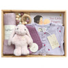 ROOM TO BLOOM Petite Plum Baby Gift Hamper set in a wooden box featuring a light pink plush bunny, a lavender baby romper, a purple muslin cloth, a wooden bead teething ring, and a book and bib.