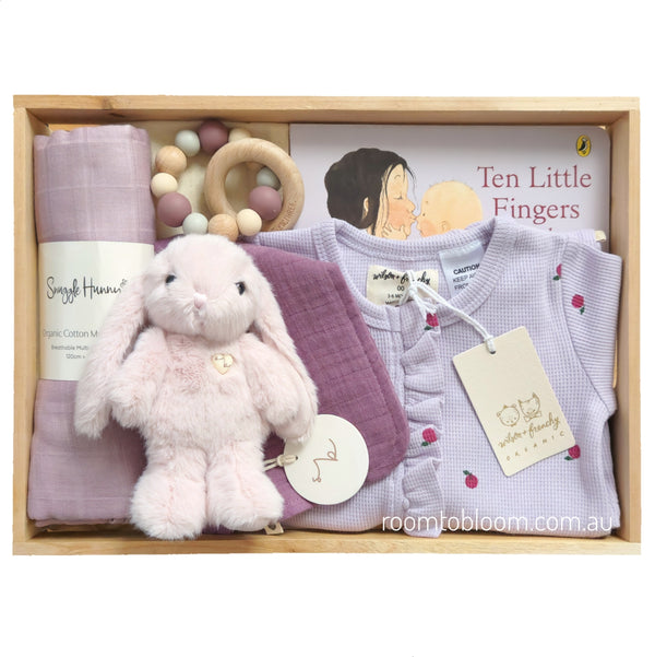 ROOM TO BLOOM Petite Plum Baby Gift Hamper set in a wooden box featuring a light pink plush bunny, a lavender baby romper, a purple muslin cloth, a wooden bead teething ring, and a book and bib.