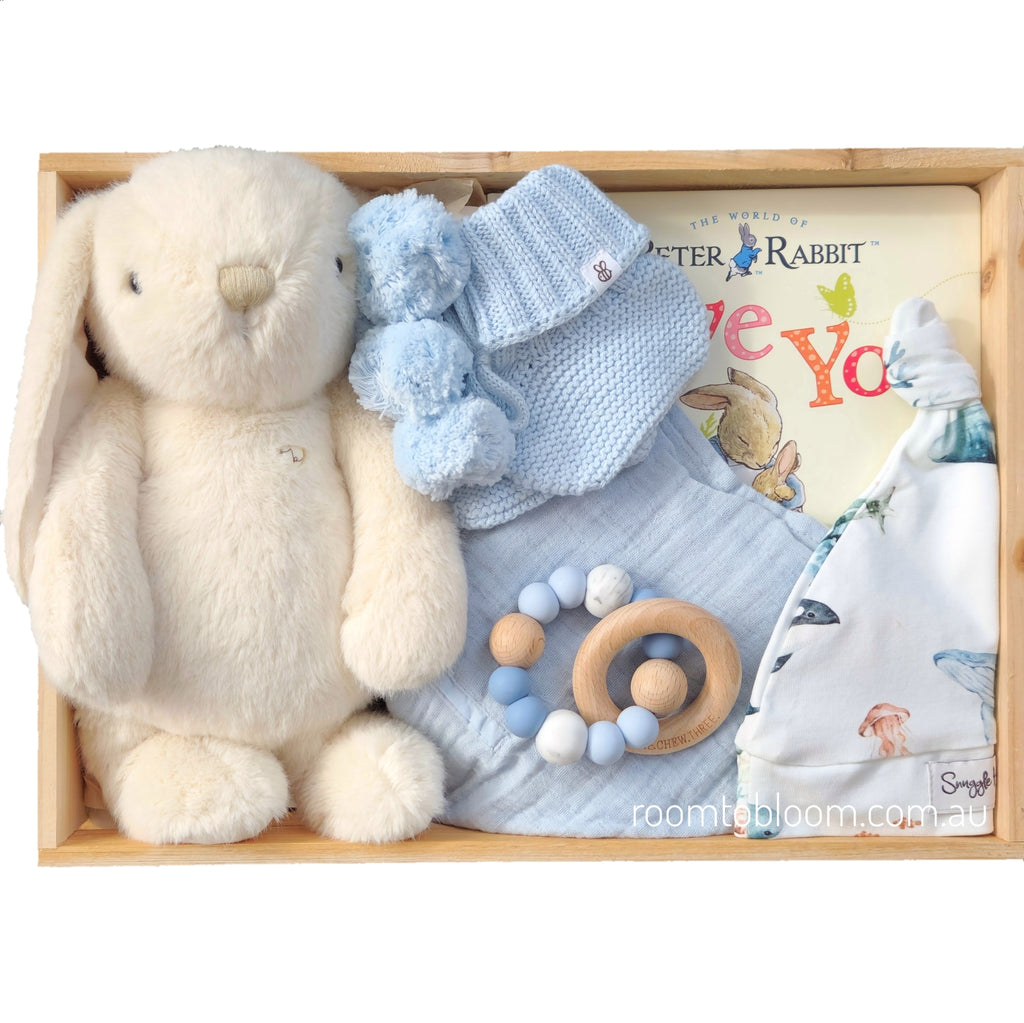 ROOM TO BLOOM Powder Blue Baby Gift Hamper set arranged in a wooden box, featuring a cream plush bunny, a light blue knit booties, a beaded teething ring, a baby book, and more.