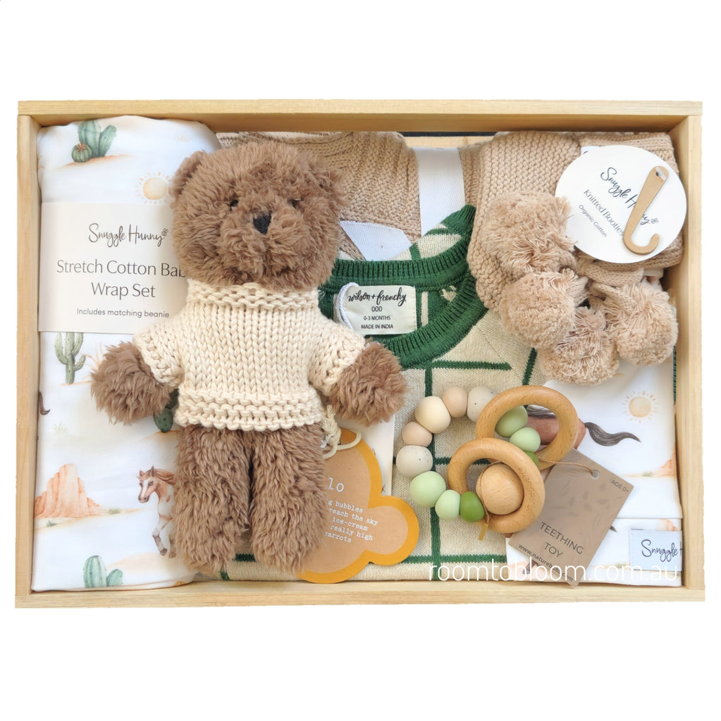 ROOM TO BLOOM Prairie Baby Gift Hamper set in a light wood box featuring a brown teddy bear wearing a cream knit sweater, a matching cotton wrap and beanie set, a green and white plaid baby growsuit, wooden and bead teething toy, and soft knit booties and blanket. 