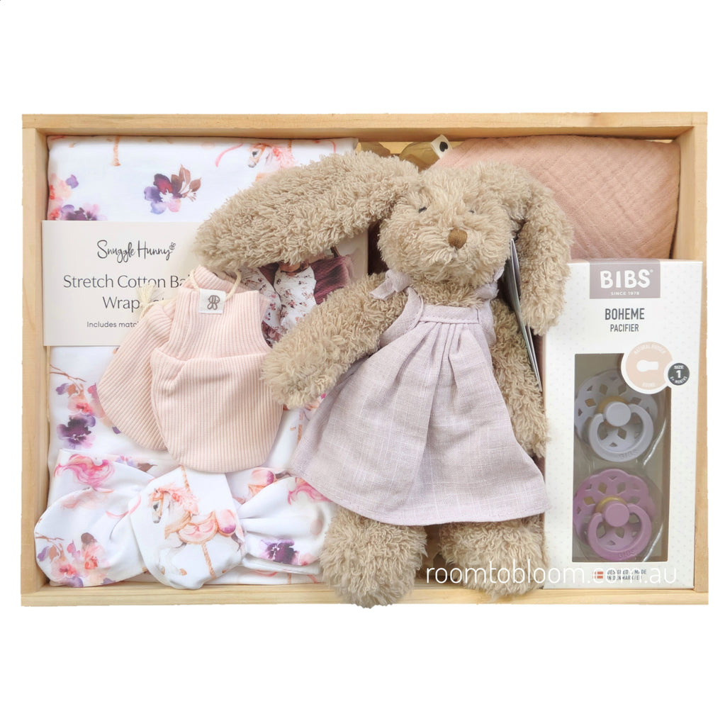 ROOM TO BLOOM Prancer Baby Gift Hamper set in a wooden box featuring a plush bunny in a pale lilac dress, pink mittens, a floral baby swaddle and headband, a pink bib, and pacifiers.