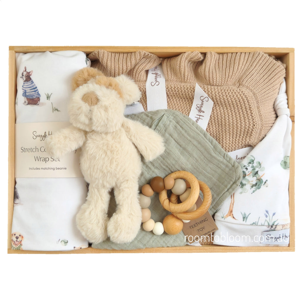 ROOM TO BLOOM Puppy Park Baby Gift Hamper set in a wooden box featuring a cream plush dog toy, a white printed wrap with a beanie, a beige knit blanket, a sage green muslin bib, and a wooden bead teether.