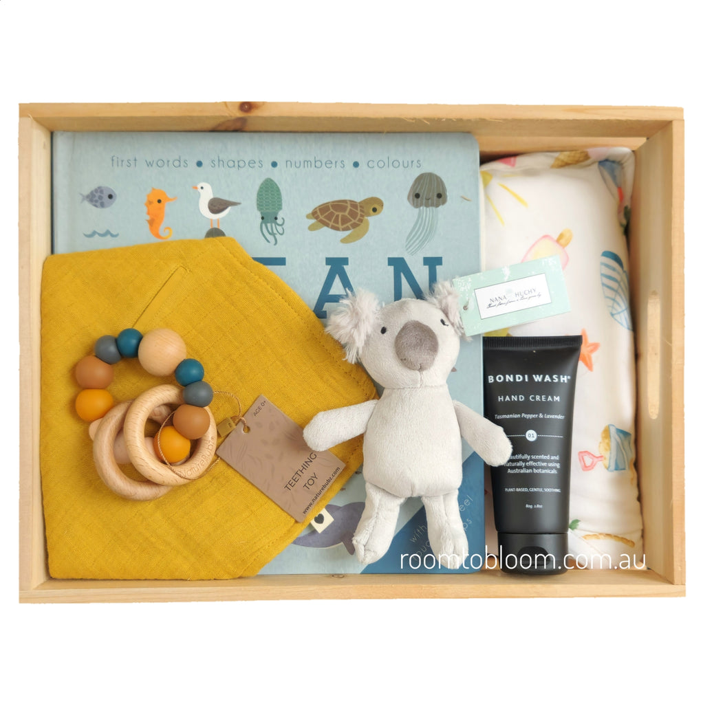 ROOM TO BLOOM Seaside Baby Gift Hamper gift box containing a baby book, a yellow muslin cloth bib, a teether with wooden rings and beads, a grey koala plush toy, a black tube of Bondi Wash hand cream, and a white printed towel.